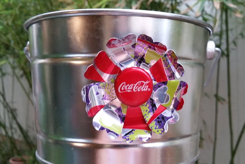 Puede incluir: Un cubo de metal plateado con una flor colorida hecha de latas de aluminio recicladas. La flor tiene un tap&oacute;n de botella de Coca-Cola rojo en el centro.