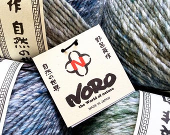 May include: Multiple skeins of variegated yarn in shades of blue, green, and grey, each wrapped with a paper band. A square white tag features the red letter N inside a black graphic, the text NORO, the world of nature, and MADE IN JAPAN.