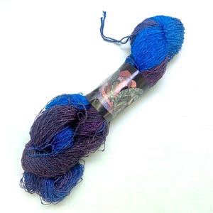 May include: A skein of yarn in shades of blue and purple, with a dark band wrapped around the center. The yarn has a textured appearance and is likely used for knitting or crocheting projects. The band features a decorative design.