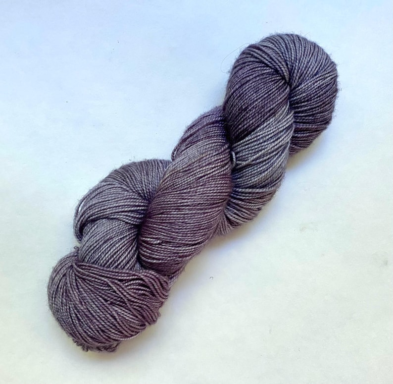 May include: A skein of yarn in shades of gray and purple, with a twisted, rope-like appearance. The yarn appears soft and is likely made of natural fibers. Ideal for knitting or crocheting projects.