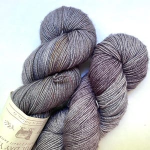 May include: Two skeins of gray yarn with a subtle color gradient. The yarn is tightly wound, showing individual strands. A label is attached to one skein, with text and a small illustration. The yarn appears soft and suitable for knitting or crochet projects.