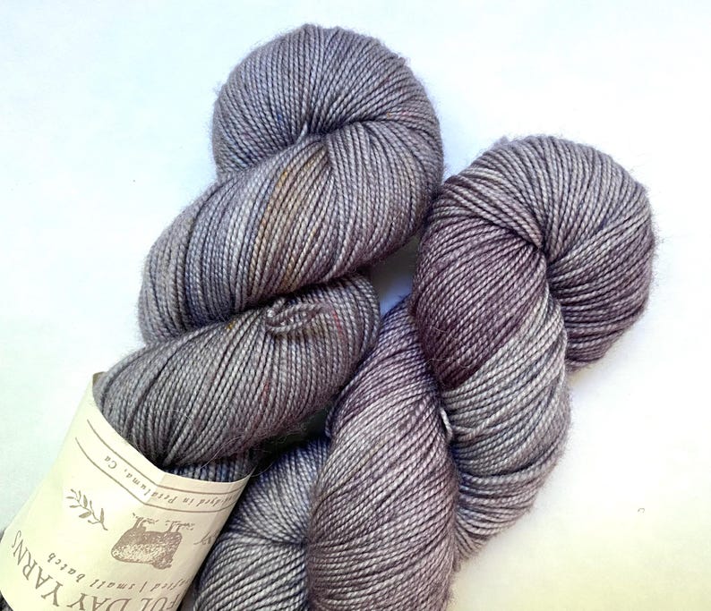 May include: Two skeins of gray yarn with a subtle color gradient. The yarn is tightly wound, showing individual strands. A label is attached to one skein, with text and a small illustration. The yarn appears soft and suitable for knitting or crochet projects.