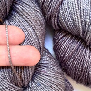 May include: Close-up of gray yarn skeins. A strand of the yarn is held between two fingers, showcasing the texture and color variations. The yarn appears soft and is suitable for knitting or crocheting projects.
