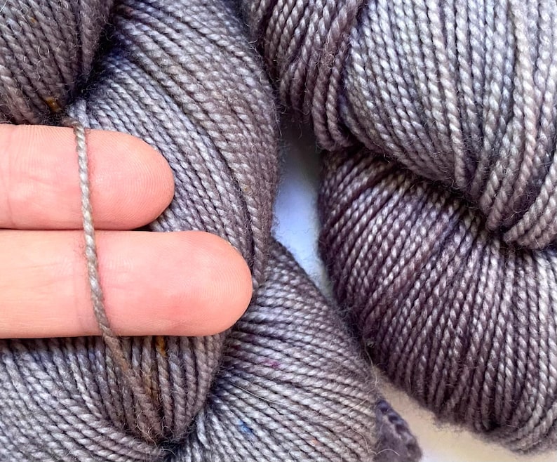 May include: Close-up of gray yarn skeins. A strand of the yarn is held between two fingers, showcasing the texture and color variations. The yarn appears soft and is suitable for knitting or crocheting projects.