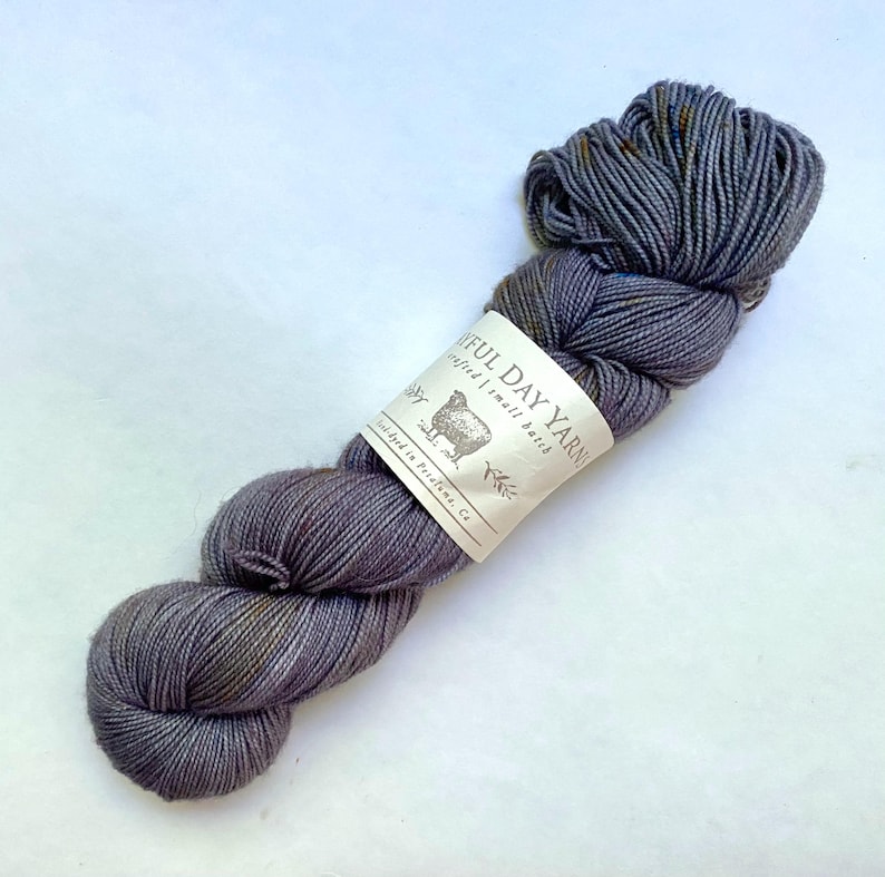 May include: A skein of gray yarn with subtle color variations and a label that reads "Joyful Day Yarn." The yarn is a soft, textured material, ideal for knitting or crocheting projects. The label features a sheep illustration.
