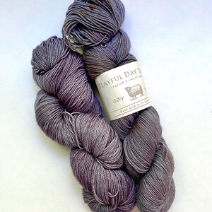 May include: Two skeins of hand-dyed yarn in shades of gray and purple. The yarn is labeled "Playful Day Yarn" with a sheep illustration. The yarn appears soft and is suitable for knitting or crochet projects.