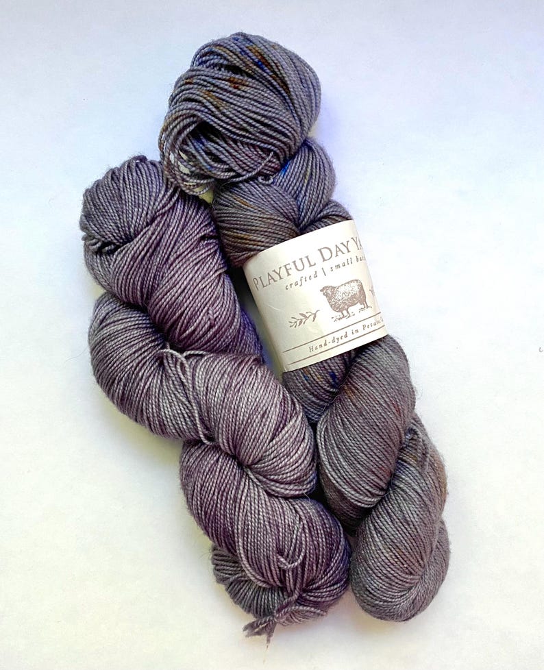 May include: Two skeins of hand-dyed yarn in shades of gray and purple. The yarn is labeled "Playful Day Yarn" with a sheep illustration. The yarn appears soft and is suitable for knitting or crochet projects.
