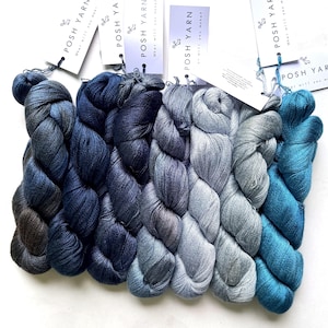 Peut inclure: Une collection de six écheveaux de fil disposés en dégradé, allant du bleu marine foncé au bleu clair et au bleu sarcelle. Chaque écheveau comporte une étiquette en papier blanc marquée POSH YARN avec le texte what will you make? attaché.
