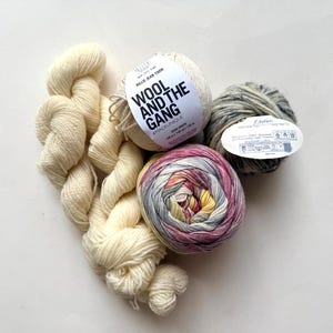 May include: An overhead shot displays a collection of yarn skeins and bundles. The assortment includes a cream-colored bundle of yarn, a white skein with the text "WOOL AND THE GANG", a gray and white skein, and a multicolored skein with pink, yellow, and blue hues.