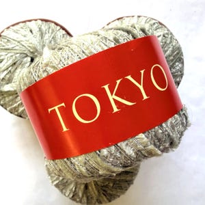 May include: A ball of metallic silver yarn wrapped in a bright red paper band with the word TOKYO printed in large, bold, gold-colored capital letters. The yarn has a shimmering, textured appearance with a reflective quality throughout the fibers.