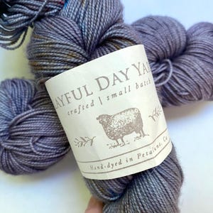 May include: A skein of hand-dyed yarn in shades of gray and blue. The yarn is labeled "Layful Day Yarn" with a sheep illustration. The label also states "crafted | small batch" and "Hand-dyed in Petaluma, Ca."