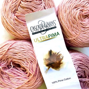 Op de afbeelding: Vier strengen garen in oudroze kleur, gerangschikt rond een wit papieren label. Het label bevat de tekst Cascade Yarns, Ultra Pima en 100% Pima Cotton, samen met een grafische illustratie van een bruine katoenbol in het midden.