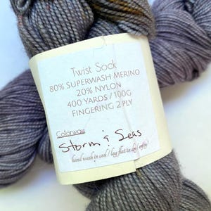 May include: A skein of gray Twist Sock yarn with a label that reads "Storm & Seas." The label also indicates the yarn is 80% Superwash Merino, 20% Nylon, 400 yards/100g, and Fingering 2 Ply. The yarn is hand-washable.