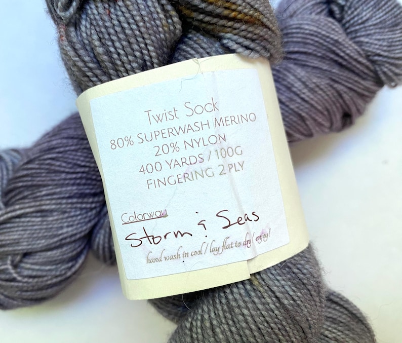 May include: A skein of gray Twist Sock yarn with a label that reads "Storm & Seas." The label also indicates the yarn is 80% Superwash Merino, 20% Nylon, 400 yards/100g, and Fingering 2 Ply. The yarn is hand-washable.