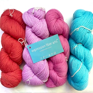 May include: A collection of colorful yarn skeins in shades of red, lavender, pink, and turquoise. A blue tag with the text "expression fiber arts" is attached to the yarn. The yarn is neatly arranged, showcasing its vibrant colors and texture.