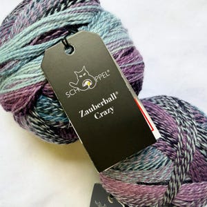 May include: A ball of variegated yarn in shades of purple, blue, and teal with a black paper tag attached. The tag features the text SCHOPPEL and Zauberball Crazy with a cat-like logo. The yarn has a twisted, multi-colored texture.