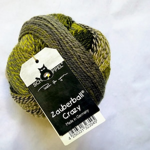 May include: A round ball of variegated yarn in shades of olive green, dark grey, and cream. A white and black paper tag is attached, featuring the text SCHOPPEL, Passion for yarn, Zauberball Crazy, Made in Germany, and a barcode.