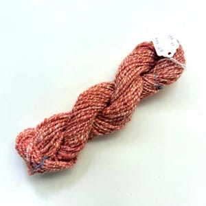 May include: A twisted skein of variegated yarn in shades of coral, orange, and white. A small white paper tag is attached to the yarn, featuring the handwritten text Art Yarn and 33 yds, indicating the length of the fiber in this specific bundle.