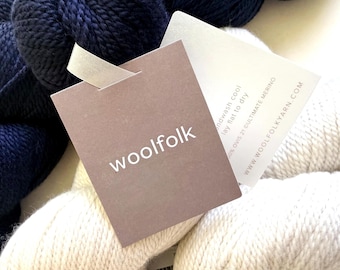 May include: Two skeins of yarn, one dark navy blue and one off-white, featuring a brown paper tag with the white text woolfolk. A secondary white tag includes the text handwash cool, lay flat to dry, 100% OVIS 21 ULTIMATE MERINO, and the website URL.