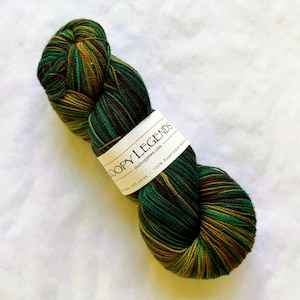 Puede incluir: Una madeja de hilo jaspeado en tonos verde bosque profundo, oliva y marrón oscuro. Una etiqueta de papel blanco envuelve el centro, impresa con el texto LOOPY LEGENDS, theloopyewe.com, 485 yards y 100% Superwash Merino.