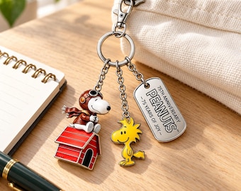 May include: A metal keychain featuring three charms: a white dog in a brown aviator cap sitting on a red doghouse, a yellow bird, and a silver rectangular tag engraved with 75TH ANNIVERSARY PEANUTS - 75 YEARS OF JOY. The charms hang from a silver ring.