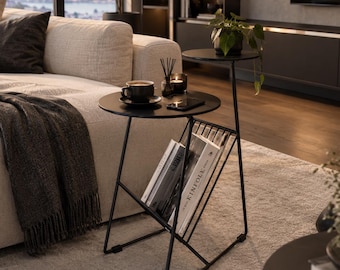 Op de afbeelding: Een zwarte metalen bijzettafel met twee ronde niveaus en een geïntegreerd tijdschriftenrek. Het onderste niveau bevat verschillende boeken, waaronder een boek met de tekst KINFLE zichtbaar op de rug. De tafel heeft een slank, hoekig frame.