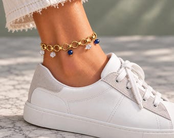 Könnte beinhalten: Ein goldfarbenes Ketten-Fußkettchen mit kreisförmigen Gliedern und baumelnden facettierten Anhängern in abwechselnd klaren und tiefblauen Farben, getragen über einem weißen Ledersneaker mit hellgrauen Wildlederakzenten.