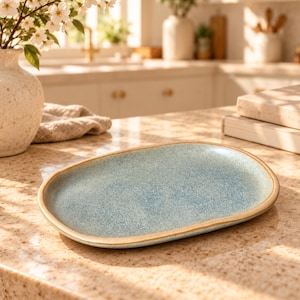 May include: An oval ceramic plate with a speckled light blue glaze and a smooth, tan-colored rim. The plate features a slightly irregular, organic shape with a shallow basin and a flat base, showcasing a textured finish throughout the surface.