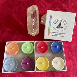 May include: A set of eight colorful tea light candles in two white boxes, a clear quartz crystal point, and a white product box labeled Balanced Being Whispering Candle Collection Four Frequency Candles 7balancedbeing.etsy.com.