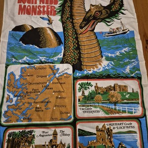 May include: A white tea towel featuring a large illustration of the Loch Ness Monster, a map of Scotland, and three framed scenes depicting The Castle Inverness, Fort Augustus The Abbey, and Urquhart Castle & Loch Ness.