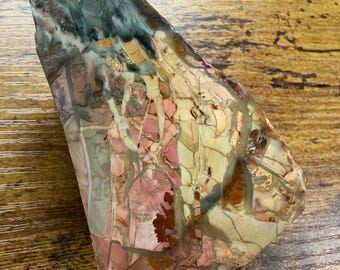 Puede incluir: Una losa pulida de piedra jaspe multicolor que presenta un patrón complejo de tonos rosa, crema, bronceado y verde salvia con vetas gris oscuro y líneas de fractura intrincadas en toda la superficie lisa, irregular y triangular.