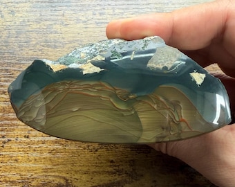 Puede incluir: Una losa de piedra ágata pulida con un borde liso y redondeado y una cara plana. La piedra muestra bandas horizontales de color tostado, beige y marrón, que hacen la transición a una sección azul grisácea translúcida con inclusiones.
