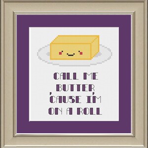 Puede incluir: Arte de punto de cruz en un marco dorado con una esterilla morada. El diseño presenta una barra de mantequilla sonriente en un plato. El texto dice "CALL ME BUTTER, 'CAUSE I'M ON A ROLL."