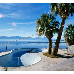 May include: A blue swimming pool with a curved design sits empty in front of a white concrete building. Two palm trees stand in the foreground, and a body of water with mountains in the distance is visible in the background.