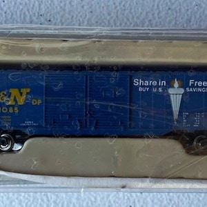 May include: A blue model train boxcar in a clear plastic display case. The car features yellow L&N lettering and a white graphic of a torch with the text Share in Freedom BUY U.S. SAVINGS BONDS printed on the side of the blue train car.