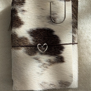 Puede incluir: Un cuaderno rectangular cubierto de piel de vaca marrón y blanca. Una fina banda elástica marrón envuelve horizontalmente el centro, con un pequeño dije de corazón de metal plateado. Un clip de metal plateado grande está sujeto en la parte superior.