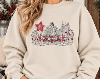May include: A beige crewneck sweatshirt featuring a graphic illustration of a Thanksgiving parade. The design includes a turkey in a top hat on a float, marching band members, a large red star, and city buildings with the text RADIO CITY.