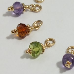 以下が含まれることがあります： 異なる色の宝石が付いた金のチャームが4つ。チャームは金のジャンプリングに取り付けられています。宝石はアメジスト、シトリン、ペリドット、アメジストです。