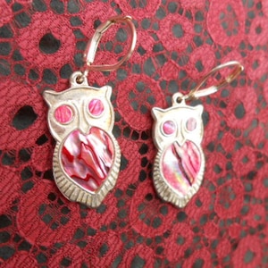 May include: A pair of silver owl earrings with pink and white iridescent shell inlays. The owls have round eyes and a heart-shaped body.