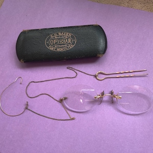 May include: A vintage pair of round, clear-framed glasses with a gold chain and a gold hair clip. The glasses are in a green leather case with the text "A.H. Bauer Manufacturing Optician 813 Houston St. Fort Worth, Tex."