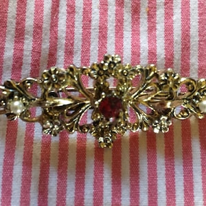 May include: A gold-tone hair clip with a red jewel in the center and two pearl accents. The clip has a delicate, ornate design.