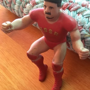 May include: A red and gold wrestling action figure with a black hat and boots. The figure is wearing a red wrestling suit with the word "USSR" printed on the front.