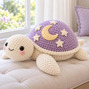 Puede incluir: Un peluche de tortuga tejido a ganchillo con cabeza y extremidades de color crema, ojos de botón negros y mejillas rosadas. La tortuga tiene un caparazón morado redondeado decorado con una luna creciente amarilla y estrellas crema.