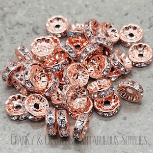 May include: A pile of rose gold-toned metal beads with clear rhinestone accents. The beads have a decorative, slightly raised design around the edge.