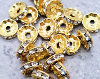 May include: A close-up of a pile of gold-toned metal bead caps with clear rhinestone accents. The bead caps are round and have a raised edge.