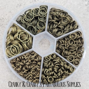 May include: A clear plastic container with six compartments filled with gold jump rings. The container is labeled "Cranky & Crabby's Fantabulous Supplies".