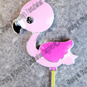 May include: Pink flamingo charm with a silver outline and a small loop at the top. The flamingo has a black beak, eye detail, and a yellow leg. The text "Cranky & Crabby's Fantabulous Supplies" is printed on the image.