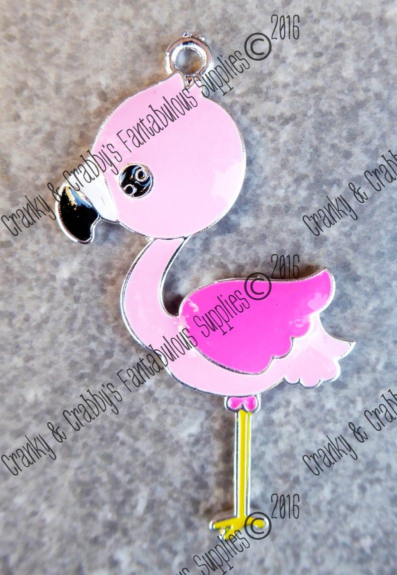 May include: Pink flamingo charm with a silver outline and a small loop at the top. The flamingo has a black beak, eye detail, and a yellow leg. The text "Cranky & Crabby's Fantabulous Supplies" is printed on the image.