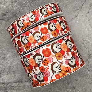 May include: Spools of ribbon with a repeating pattern of a white-masked face, orange pumpkins, and flowers in shades of orange, red, and pink on a cream background. The ribbon is suitable for Halloween crafts.