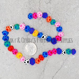 Op de afbeelding: Een streng met kleurrijke poot afdruk kralen. De kralen zijn gemaakt van polymer clay en zijn ongeveer 1,2 cm in diameter. De kralen zijn geregen aan een doorzichtig elastisch koord. De kralen zijn in verschillende kleuren, waaronder roze, blauw, groen, geel, oranje, rood en paars. Sommige kralen hebben zwarte en witte stippen.